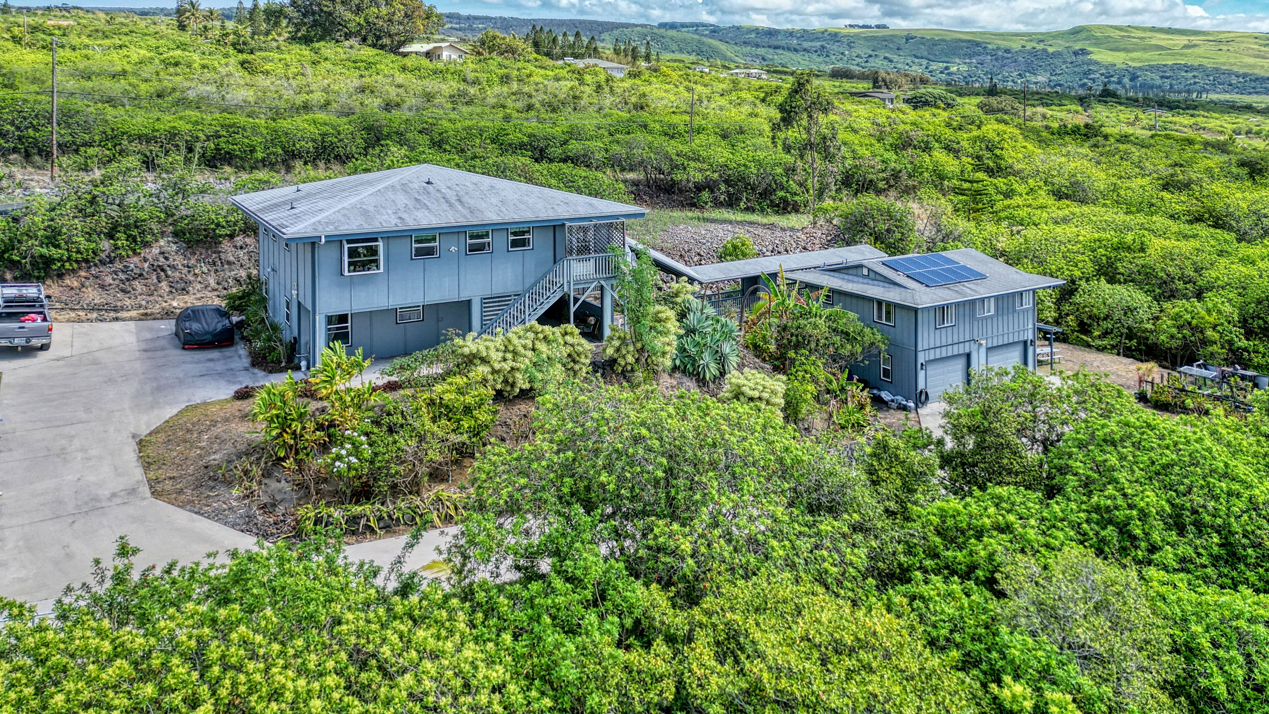 Aerial view of the spacious two-story Big Island home with ohana over a 2-car garage, surrounded by lush tropical landscaping and set against sweeping green countryside and ocean views.