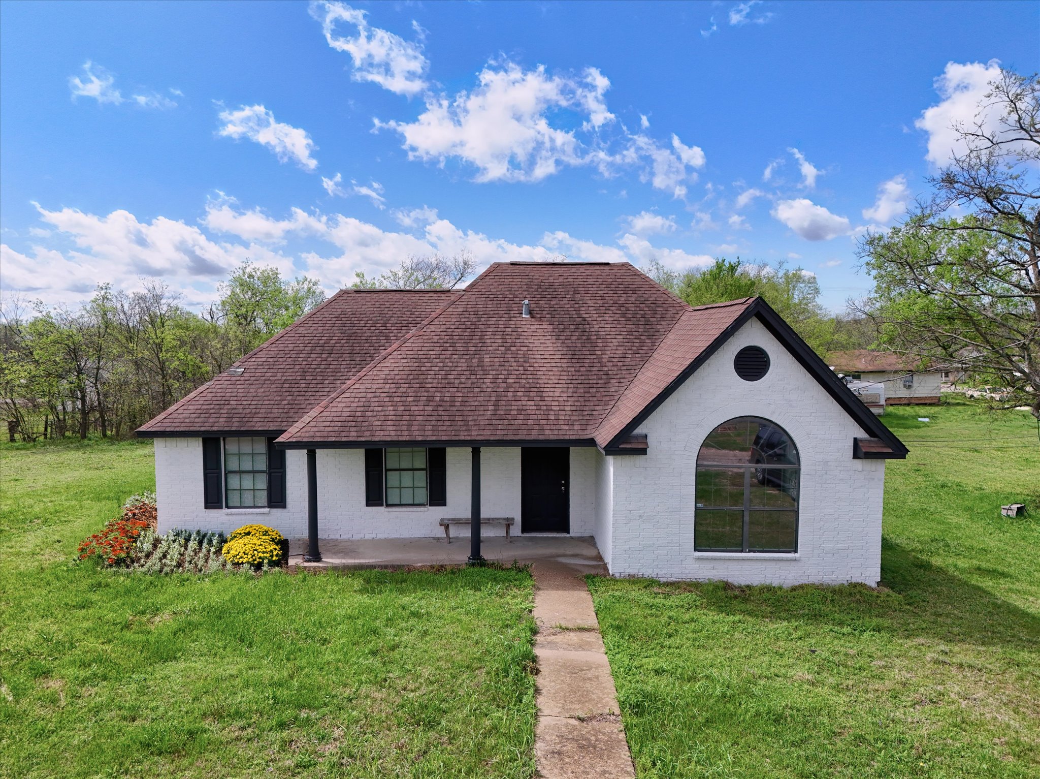 Single story home featuring a shingled roof, covered porch, a front yard, and brick siding. Flowers added using AI